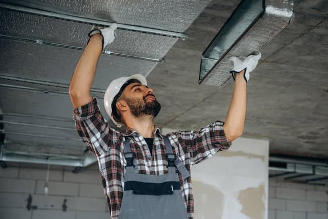 A bearded technician wearing a white hard hat, plaid shirt, and gray overalls reaches up to install or adjust metal ductwork in an exposed ceiling.