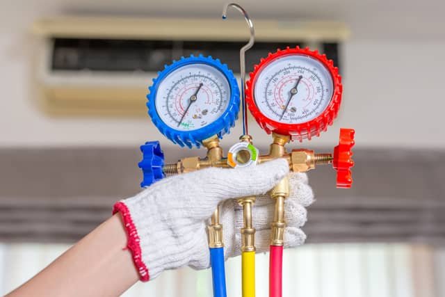 A gloved hand holding a set of red and blue refrigerant manifold gauges with colored hoses, with an air conditioning unit visible in the background.