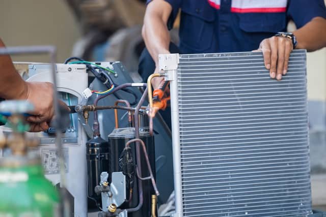 Two technicians working on an air conditioning unit, with one holding copper refrigerant lines while the other steadies the condenser coil panel.