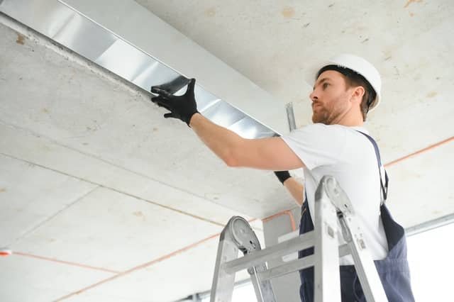 A technician wearing a white hard hat and work gloves stands on a ladder while installing rectangular metal ductwork along a ceiling in an unfinished space.