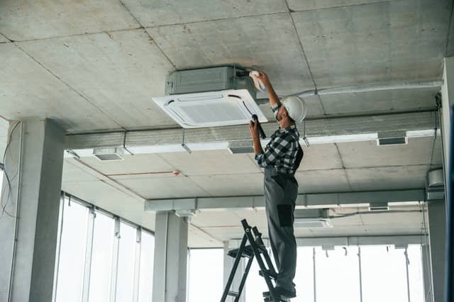 A technician standing on a ladder installing or servicing a ceiling-mounted cassette air conditioning unit in a commercial space with exposed concrete ceilings.