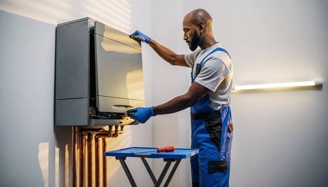 A technician wearing blue overalls and protective gloves installs or services a gray wall-mounted boiler unit with copper piping below it.