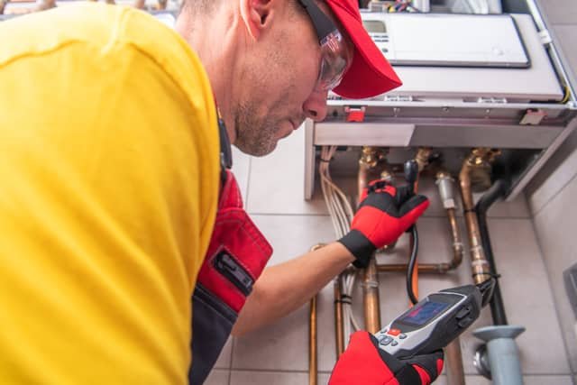 A technician in a yellow shirt and red cap uses a digital diagnostic meter while inspecting copper pipes and valves on a heating system.
