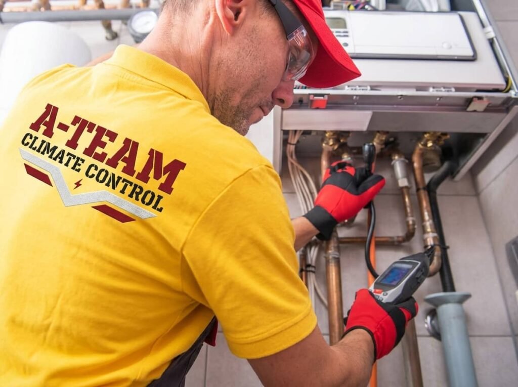 A technician wearing a yellow shirt and red cap uses a handheld diagnostic meter to inspect copper piping connected to a wall-mounted heating system.