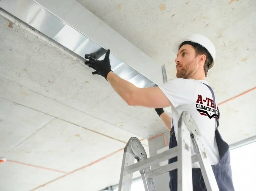 A technician wearing a white hard hat and company t-shirt stands on a ladder while installing metal ductwork near the ceiling of an unfinished space.