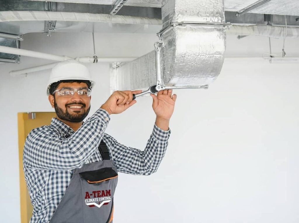A smiling HVAC technician wearing a hard hat and safety glasses uses a ratchet wrench to secure insulated ductwork mounted to a ceiling.