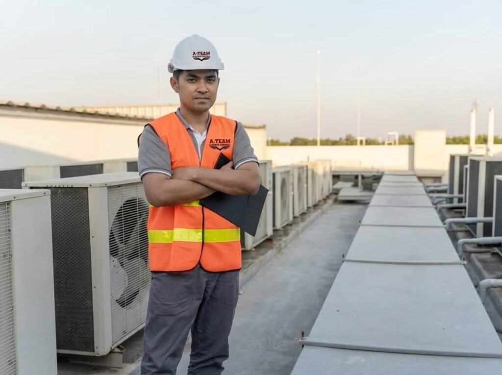A technician wearing an orange safety vest and white hard hat stands with arms crossed on a commercial building rooftop lined with air conditioning units.